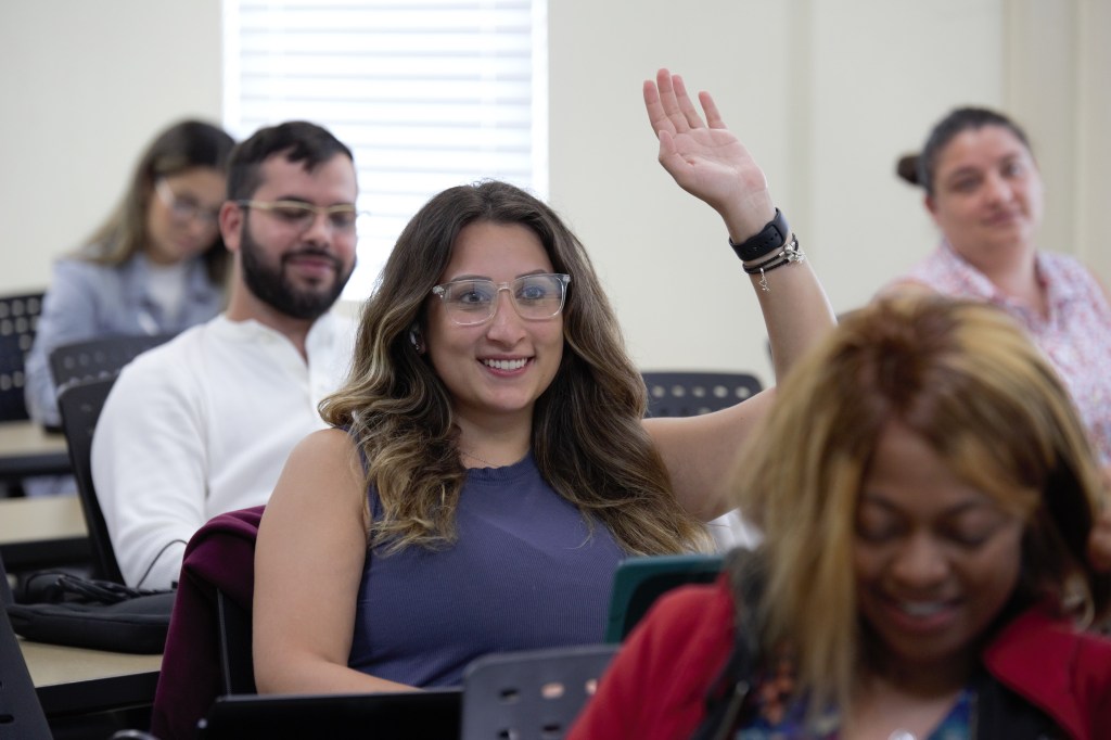 A woman in glasses raising her hand to contribute her thoughts in MUA classroom.